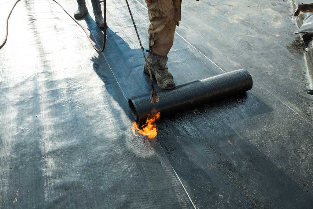 Workers secure waterproof covering around a roof opening while lumber and tools remain on a protected work platform.