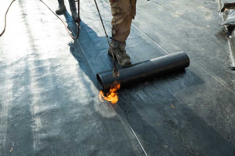 Workers secure waterproof covering around a roof opening while lumber and tools remain on a protected work platform.