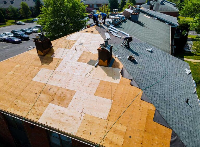 Workers remove old shingles from a house roof while exposed decking and protective ground coverings are visible.